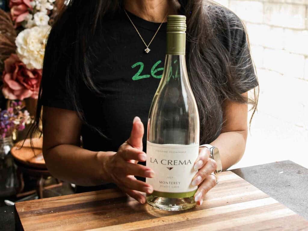 A woman in a black shirt holds an empty La Crema Monterey Pinot Gris wine bottle on a wooden cutting board, with flowers and a window in the background.